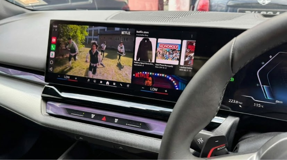 Interieur van een moderne auto met een breed panoramisch dashboard-scherm dat een split-screen weergave toont, met links een video die afspeelt en rechts de Netflix-interface.